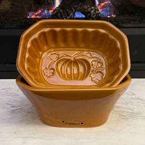 May include: Two-piece, pumpkin-shaped ceramic baking dish set in a warm, orange-brown hue. The top dish features a pumpkin and leaf design. The bottom dish is a slightly larger, complementary shape. The brand name "Crate & Barrel" is visible.