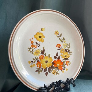May include: A white ceramic dinner plate with a brown rim and a floral design featuring yellow, orange, and brown flowers.
