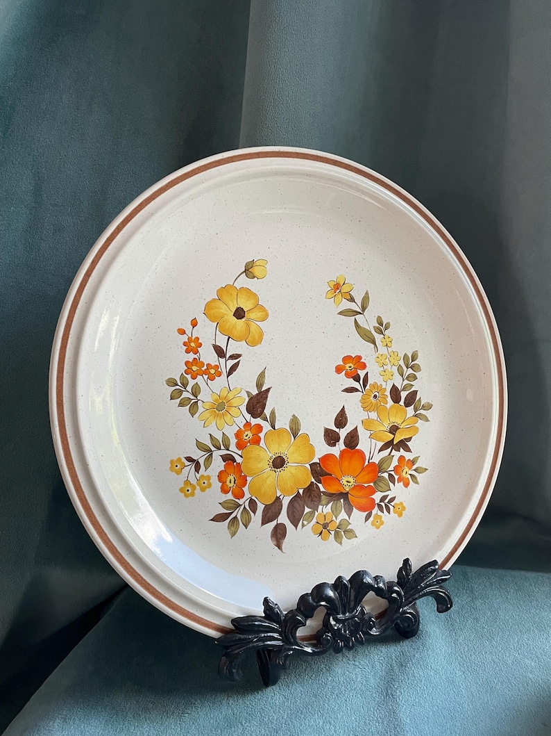 May include: A white ceramic dinner plate with a brown rim and a floral design featuring yellow, orange, and brown flowers.