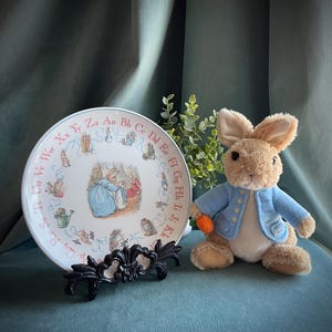May include: A white ceramic plate with a Peter Rabbit alphabet design. The plate features a scene of Peter Rabbit and his mother in the garden. The plate is on a black stand. A plush Peter Rabbit toy is sitting next to the plate.