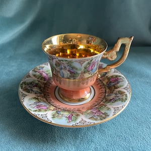May include: A vintage teacup and saucer set with a gold rim and intricate floral designs. The cup has a gold interior and the saucer features a scene of people in a garden.