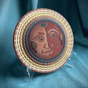 May include: A circular ceramic wall plaque featuring a central relief of a dual-faced sun and moon design in shades of terracotta and dark brown. The outer border displays a radiating pattern of white, yellow, and orange textured segments.