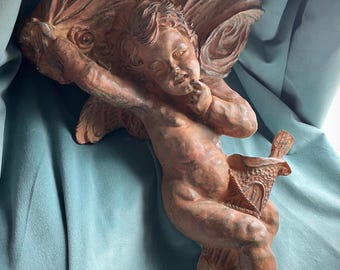 May include: A terracotta-colored cherub sculpture with detailed wings and a basket. The cherub is in a seated position, with one hand near its mouth. The sculpture has a classical design and is set against a blue background.