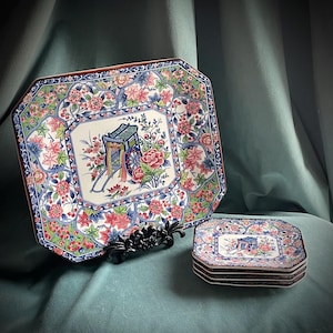 May include: A large, octagonal decorative plate and a stack of smaller plates. The plates feature a floral design with pink, blue, and green accents. The center of each plate has a detailed illustration of a cart with flowers.