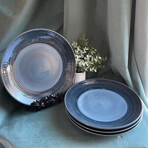 May include: A set of four blue ceramic plates with a textured rim and a decorative border. One plate is displayed on a black decorative stand. The plates have a light blue center and a darker blue rim.