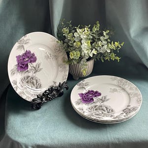 May include: White ceramic plates with a purple floral design. The plates feature a large purple flower and gray floral accents. A black decorative stand and a small potted plant are also visible.