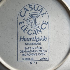 May include: A white ceramic plate with a blue and white logo that reads "Casual Elegance Hearthside Stoneware". The logo features a wine glass and grapes. The plate also has the text "Safe in your dishwasher, oven, & microwave oven Chablis Japan 571" printed on the bottom.