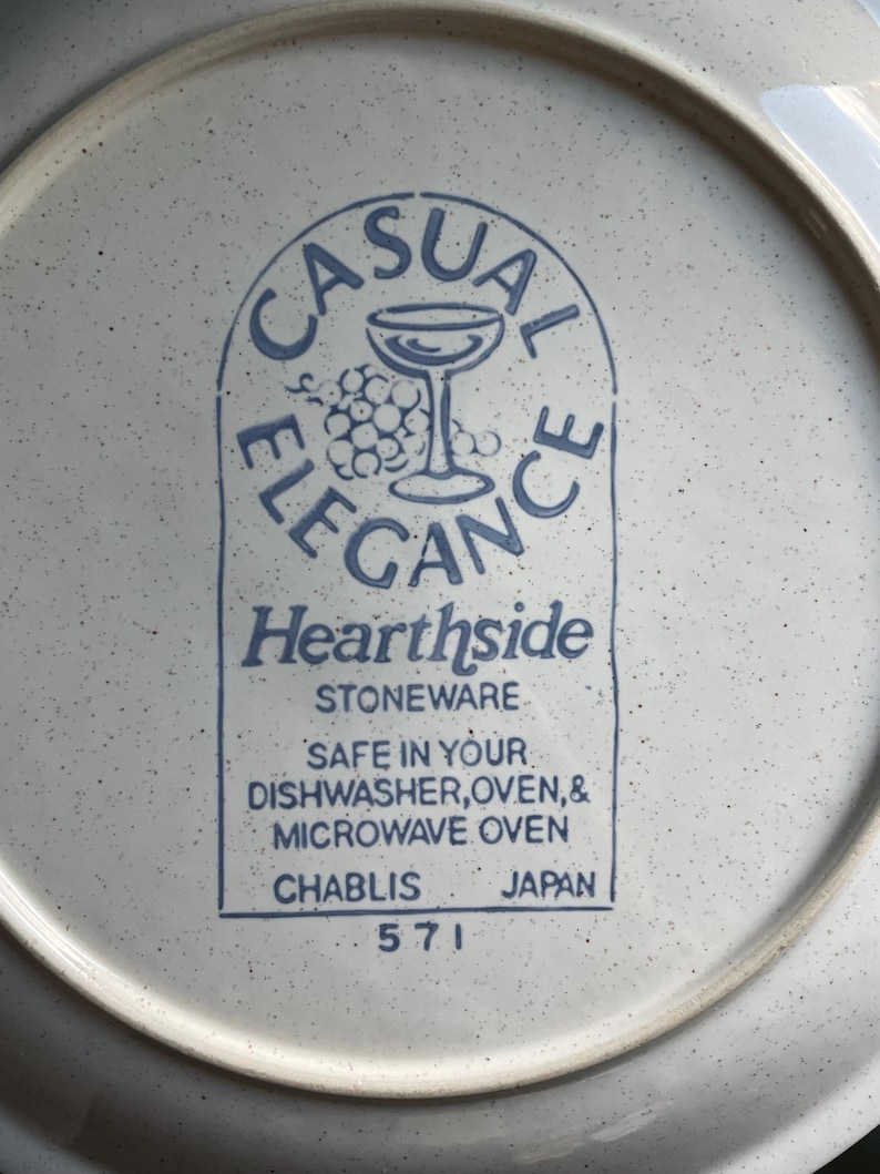 May include: A white ceramic plate with a blue and white logo that reads "Casual Elegance Hearthside Stoneware". The logo features a wine glass and grapes. The plate also has the text "Safe in your dishwasher, oven, & microwave oven Chablis Japan 571" printed on the bottom.