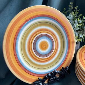 May include: A set of colorful ceramic plates with concentric circles in shades of orange, yellow, blue, and white. The plates are displayed on a black decorative stand, with a stack of additional plates visible in the background. The plates have a rustic, handcrafted appearance.