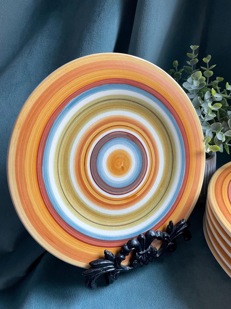 May include: A set of colorful ceramic plates with concentric circles in shades of orange, yellow, blue, and white. The plates are displayed on a black decorative stand, with a stack of additional plates visible in the background. The plates have a rustic, handcrafted appearance.