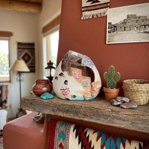 May include: A hollow, teardrop-shaped ceramic sculpture featuring a hand-painted scene of a person in a pink skirt standing near a tan brick structure, a tree, and a blue water feature, with a large cutout opening on the upper front section.