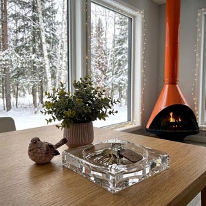 May include: A clear square acrylic tray holding a set of metal keys with a brown leather tag, a small ceramic bird figurine with a speckled finish, and a potted green plant in a textured pinkish-beige pot on a light wood table surface.
