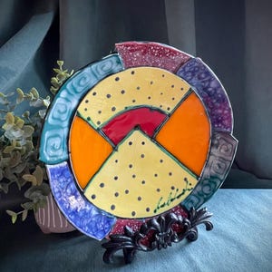 May include: A colorful decorative plate with a unique geometric design. The plate features a yellow center with black dots, surrounded by segments of orange, red, blue, and purple. It rests on a black metal stand with ornate detailing.