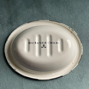 May include: An oval, white soap dish with a black rim. The dish has a ridged interior and the words "MACKENZIE-CHILDS" are printed in black, along with a small emblem. The dish is set against a teal background.