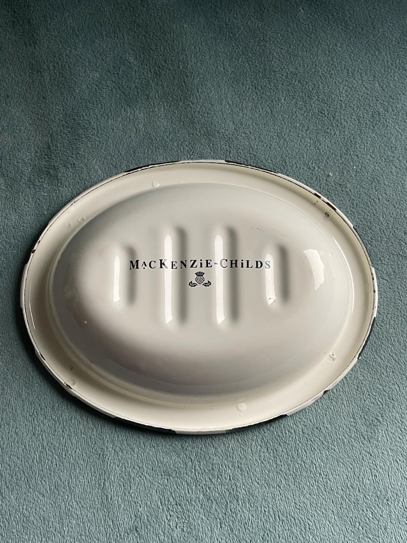 May include: An oval, white soap dish with a black rim. The dish has a ridged interior and the words "MACKENZIE-CHILDS" are printed in black, along with a small emblem. The dish is set against a teal background.