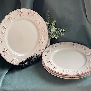 May include: Off-white ceramic plates with embossed seashell and starfish designs around the rims. The plates have a slightly distressed, rustic look. One plate is displayed on a black metal stand, and three plates are stacked.