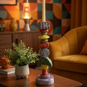 May include: A tall, colorful ceramic candle holder with a lit cream-colored candle. The holder is composed of stacked, textured, geometric shapes in shades of gray, red, yellow, and green. The candle holder sits on a wooden table.