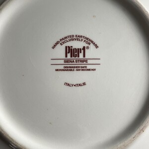May include: A close-up of the underside of a white plate. The plate features a brown logo with the text "Pier 1 Siena Stripe". The text "Hand-Painted Earthenware Exclusively For" is above the logo. The plate is dishwasher and microwave safe.