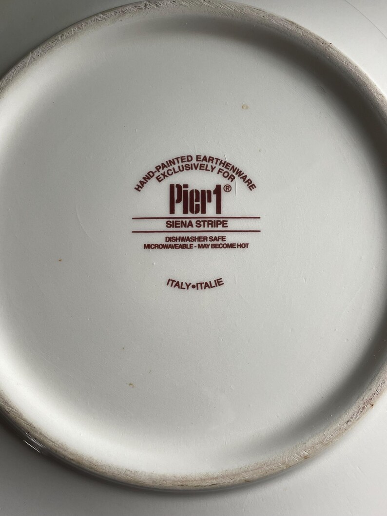 May include: A close-up of the underside of a white plate. The plate features a brown logo with the text "Pier 1 Siena Stripe". The text "Hand-Painted Earthenware Exclusively For" is above the logo. The plate is dishwasher and microwave safe.