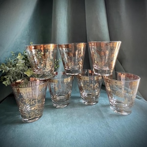 May include: Set of seven vintage clear drinking glasses with gold rims and ornate gold floral designs. The glasses are arranged in a semi-circle on a teal surface, with a green plant in the background. The glasses are of varying heights.