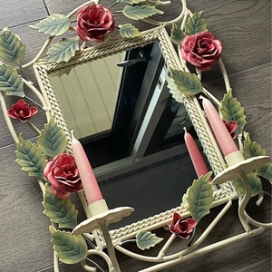 May include: White metal mirror with a decorative floral design featuring pink roses and leaves. The mirror has two candle holders with pink candles.