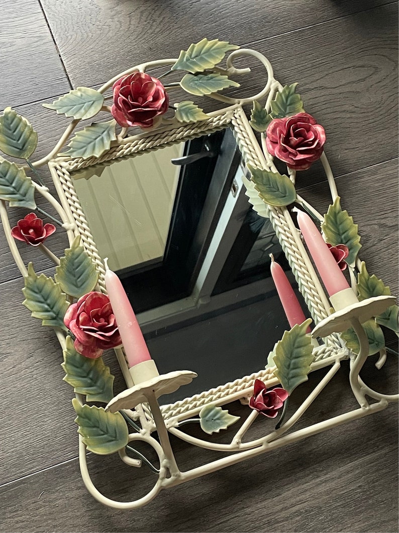 May include: White metal mirror with a decorative floral design featuring pink roses and leaves. The mirror has two candle holders with pink candles.