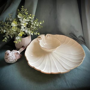 May include: A cream-colored, shell-shaped serving dish with a smaller, matching bowl nested inside. The dish has a gold trim and a ribbed design. A decorative bird figurine and a small potted plant are also present.