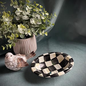 May include: A decorative arrangement featuring a ceramic bird figurine with brown and white details, a black and white checkered oval bowl, and a potted plant with green foliage. The items are arranged on a teal surface.