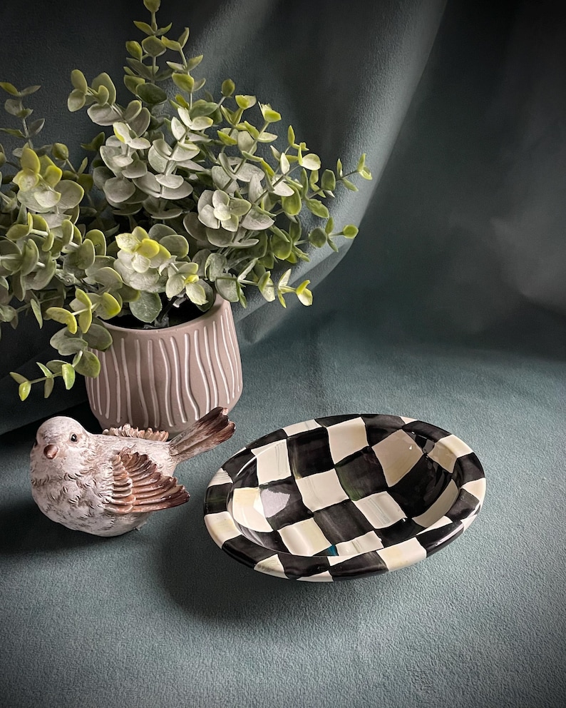 May include: A decorative arrangement featuring a ceramic bird figurine with brown and white details, a black and white checkered oval bowl, and a potted plant with green foliage. The items are arranged on a teal surface.