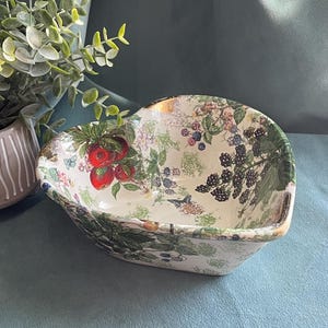 May include: A heart-shaped ceramic bowl with a botanical design featuring red apples, blackberries, and floral accents. The bowl has a gold rim and is set against a blue background.