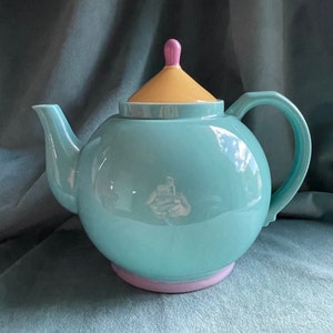 May include: A teal blue teapot with a yellow lid and a pink knob. The teapot has a rounded body and a spout on one side and a handle on the other.