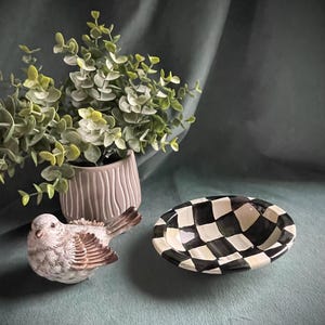 May include: A decorative arrangement featuring a ceramic bird figurine, a small checkered bowl, and a potted plant with green foliage. The bowl has a black and white checkerboard pattern. The bird is white and brown.