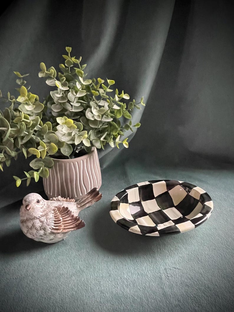 May include: A decorative arrangement featuring a ceramic bird figurine, a small checkered bowl, and a potted plant with green foliage. The bowl has a black and white checkerboard pattern. The bird is white and brown.