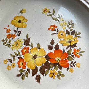 May include: A white ceramic dinner plate with a brown rim and a floral design featuring yellow, orange, and brown flowers.