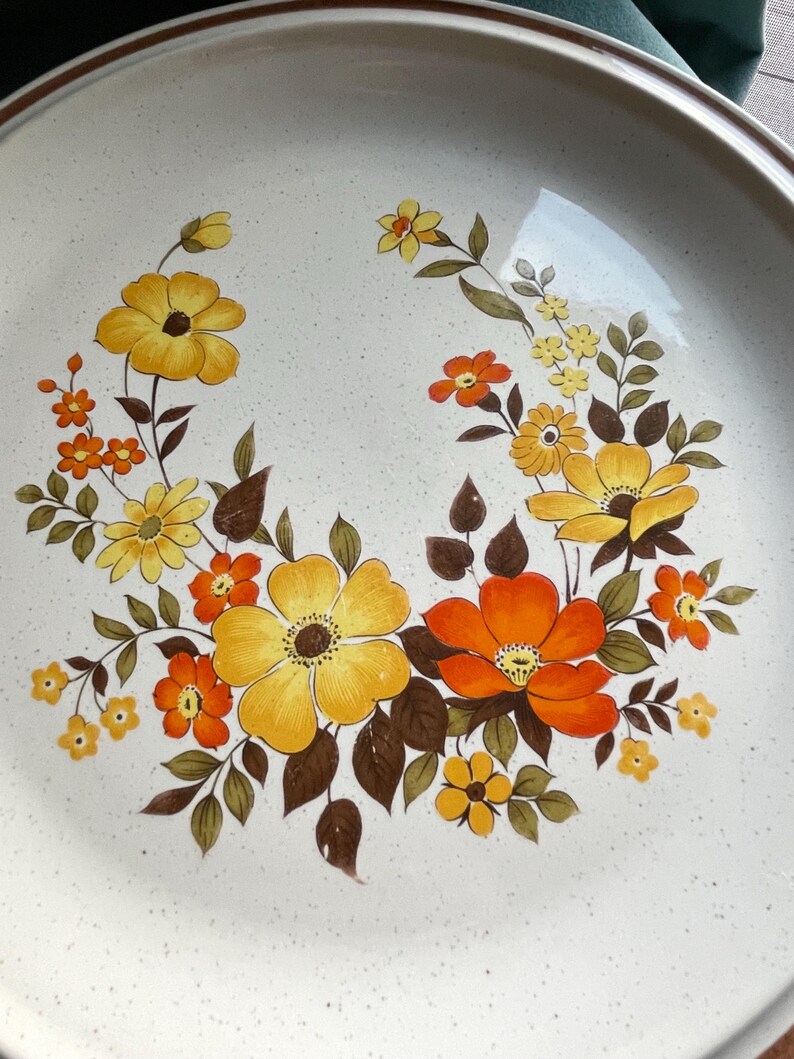 May include: A white ceramic dinner plate with a brown rim and a floral design featuring yellow, orange, and brown flowers.