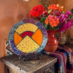May include: A round ceramic decorative plate featuring a vibrant geometric design with yellow sections containing black dots, solid orange segments, and a red arc. The plate has a dark purple and teal patterned border and rests on a black metal stand.