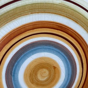 May include: Close-up of a ceramic plate with a spiral design. The plate features concentric rings of color, including brown, blue, tan, and white. The surface has a crackle glaze effect, adding texture to the design. The plate is a decorative item.