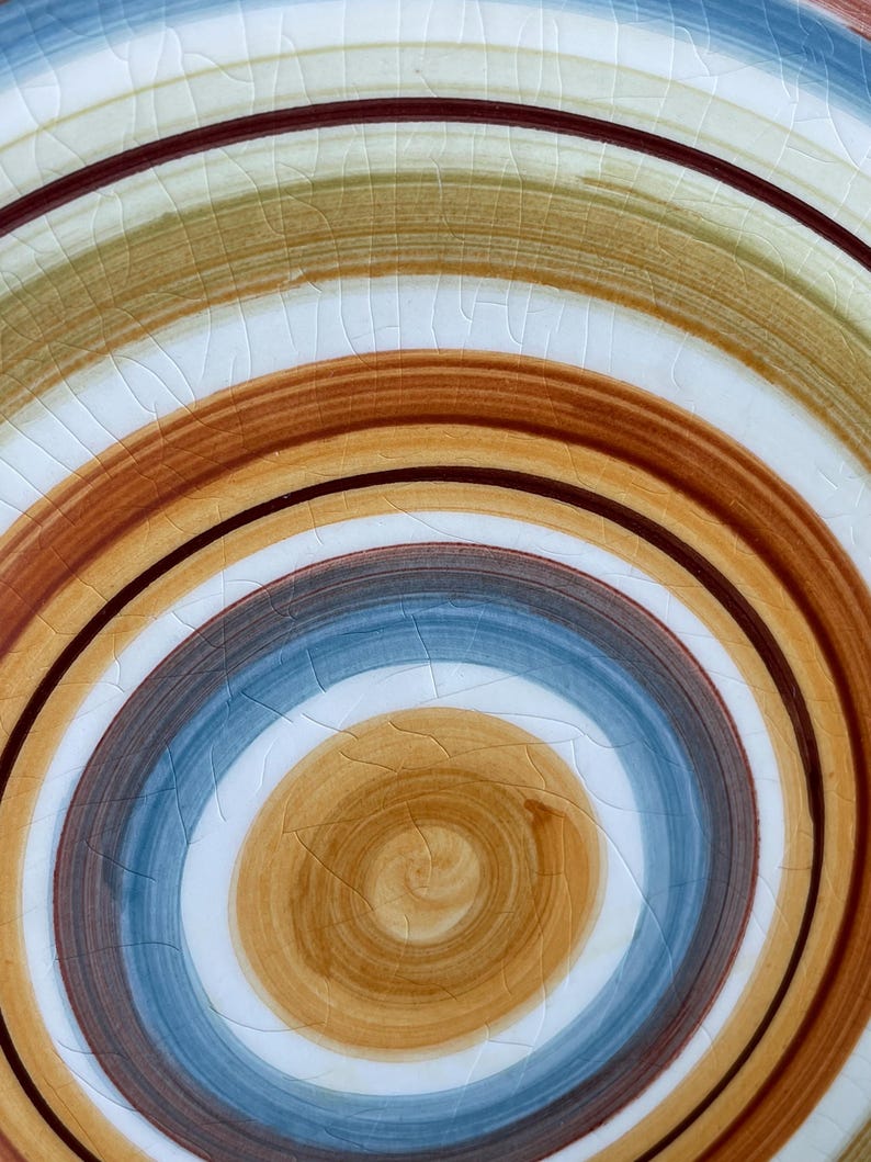 May include: Close-up of a ceramic plate with a spiral design. The plate features concentric rings of color, including brown, blue, tan, and white. The surface has a crackle glaze effect, adding texture to the design. The plate is a decorative item.