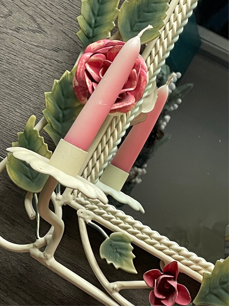 May include: White metal wall mirror with a floral design. The mirror has a rope-like border and two pink candlesticks. The candlesticks are decorated with pink roses.