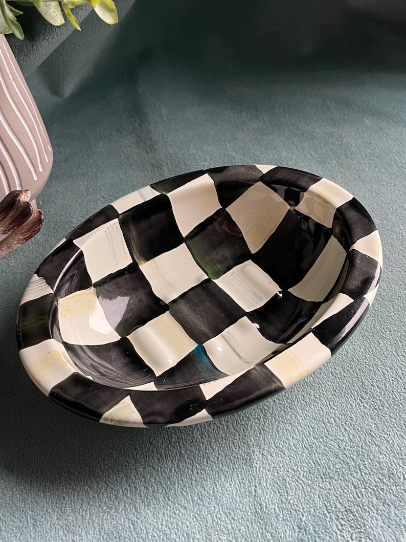 May include: Oval ceramic dish featuring a black and white chequered design. The dish has a shallow, curved interior and a glossy finish. The rim is painted cream. This decorative dish is suitable for holding small items, such as soap.