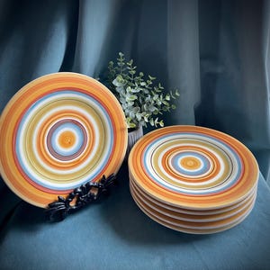 May include: A set of six ceramic plates with concentric circles in orange, yellow, blue, and white. The plates are stacked on a teal surface, with one plate displayed on a decorative black stand. A small plant is in the background.