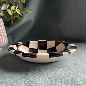 May include: A small, white ceramic bowl with a black and white chequered rim. The bowl has a shallow design and a smooth, glossy finish. The bowl is likely for serving snacks or small portions.