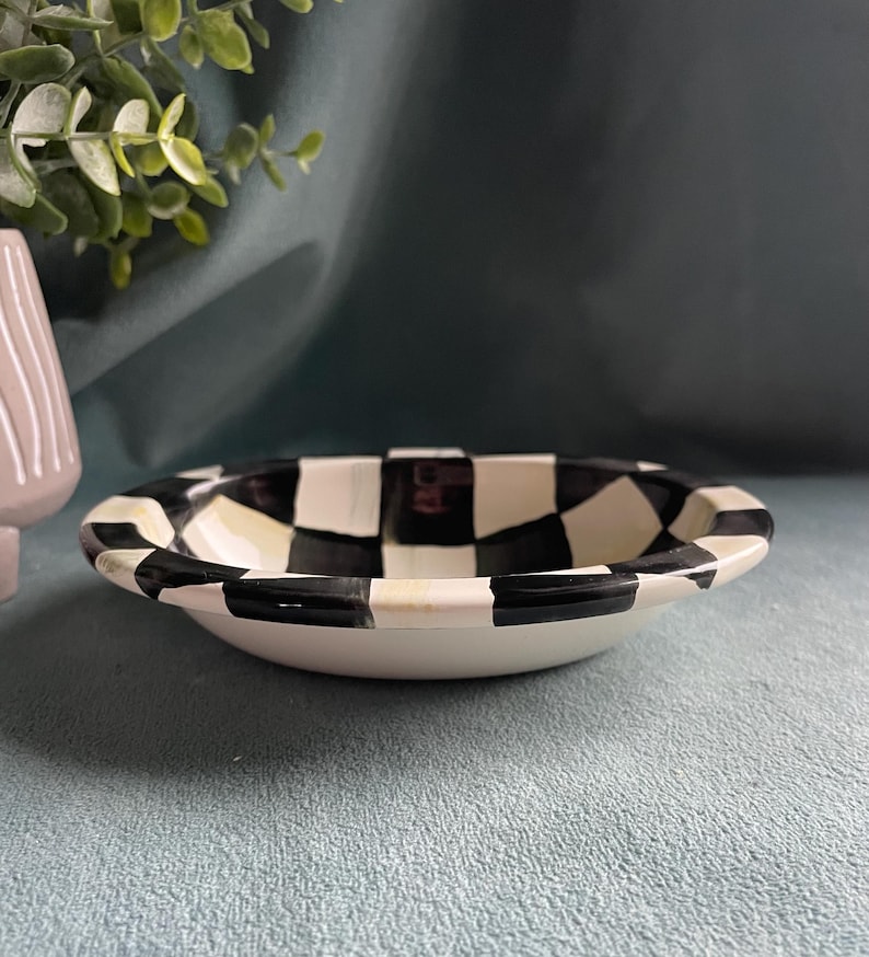 May include: A small, white ceramic bowl with a black and white chequered rim. The bowl has a shallow design and a smooth, glossy finish. The bowl is likely for serving snacks or small portions.