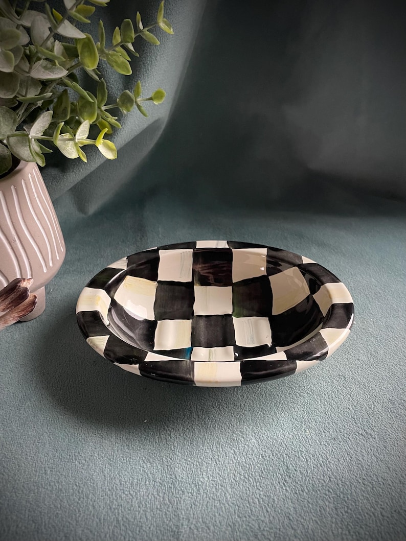May include: An oval ceramic bowl featuring a black and white chequered design. The bowl has a glossy finish and a raised rim. The interior is plain white. This bowl is likely for serving or display purposes. The bowl's dimensions are approximately 20 cm in length.