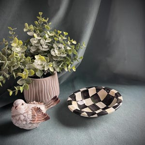 May include: A ceramic bird figurine with brown and white feathers sits near a small, oval black and white chequered dish. A potted plant with green leaves is in the background.
