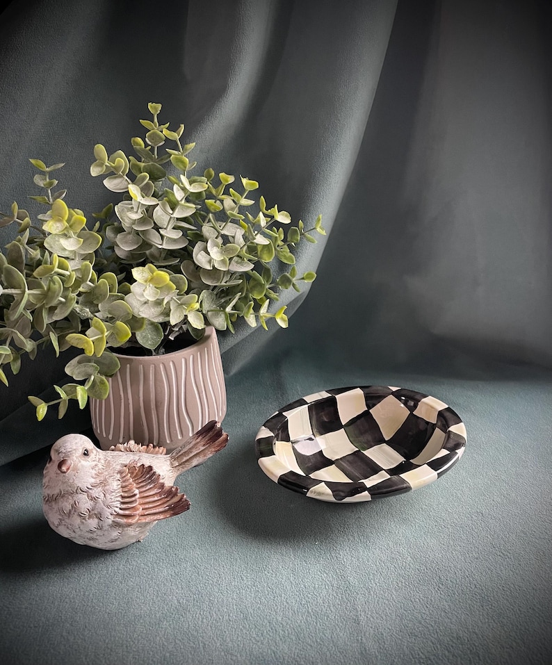 May include: A ceramic bird figurine with brown and white feathers sits near a small, oval black and white chequered dish. A potted plant with green leaves is in the background.