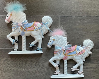 May include: Two white ceramic carousel horse figurines. Each horse has a pink and blue saddle, a blue and pink feathered mane, and decorative bows. The horses are mounted on a white base.