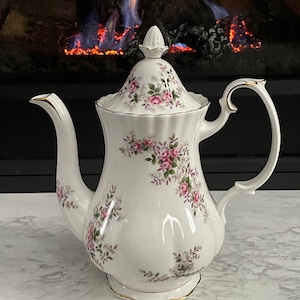 May include: A white porcelain teapot with a floral pattern of pink roses and green leaves. The teapot has a gold trim around the lid and base.