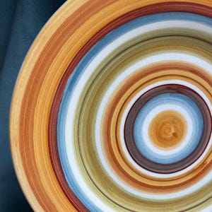 May include: A ceramic plate with a spiral design. The plate features concentric bands of color, including shades of orange, brown, blue, green, and white. The design is centered, creating a visually appealing pattern. The plate has a slightly cracked surface.