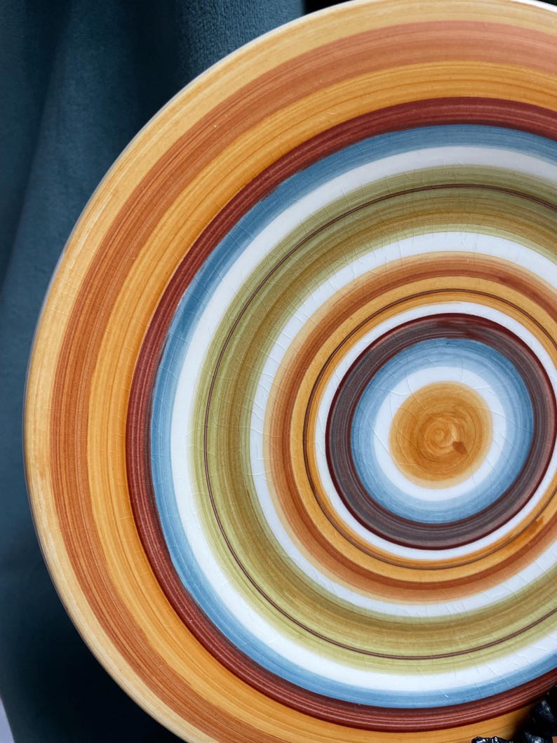 May include: A ceramic plate with a spiral design. The plate features concentric bands of color, including shades of orange, brown, blue, green, and white. The design is centered, creating a visually appealing pattern. The plate has a slightly cracked surface.
